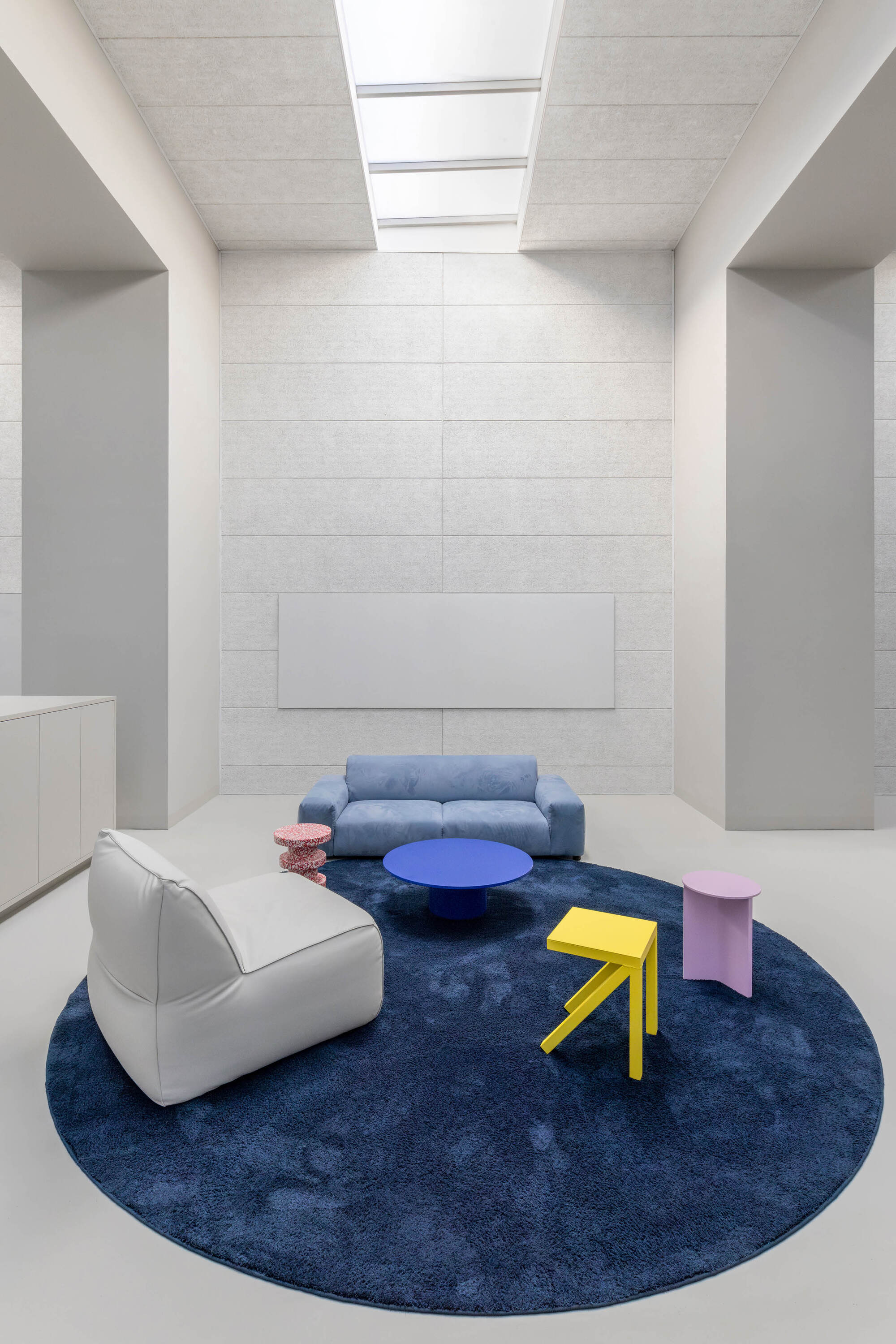 Lounge corner with a blue sofa, white armchair and yellow stool on a round navy rug beneath a skylight