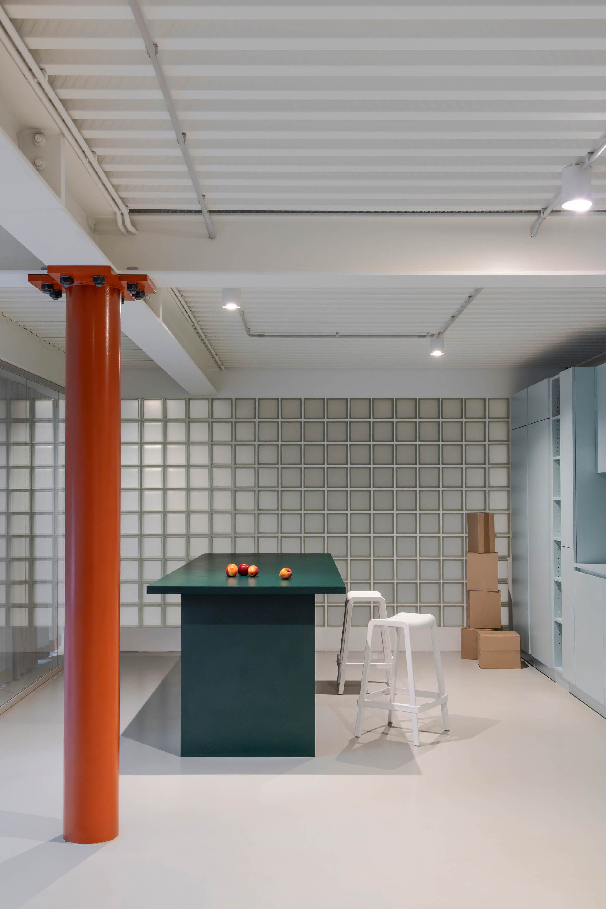 Green kitchen island with three apples beside an orange steel column, backed by a glass block wall and stacked cardboard boxes