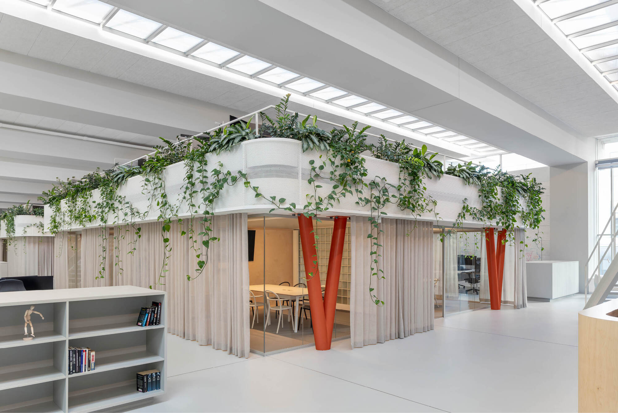 Raised white planter volume on V-shaped orange columns, with pink curtains and a glazed meeting room below, lit by rooflights