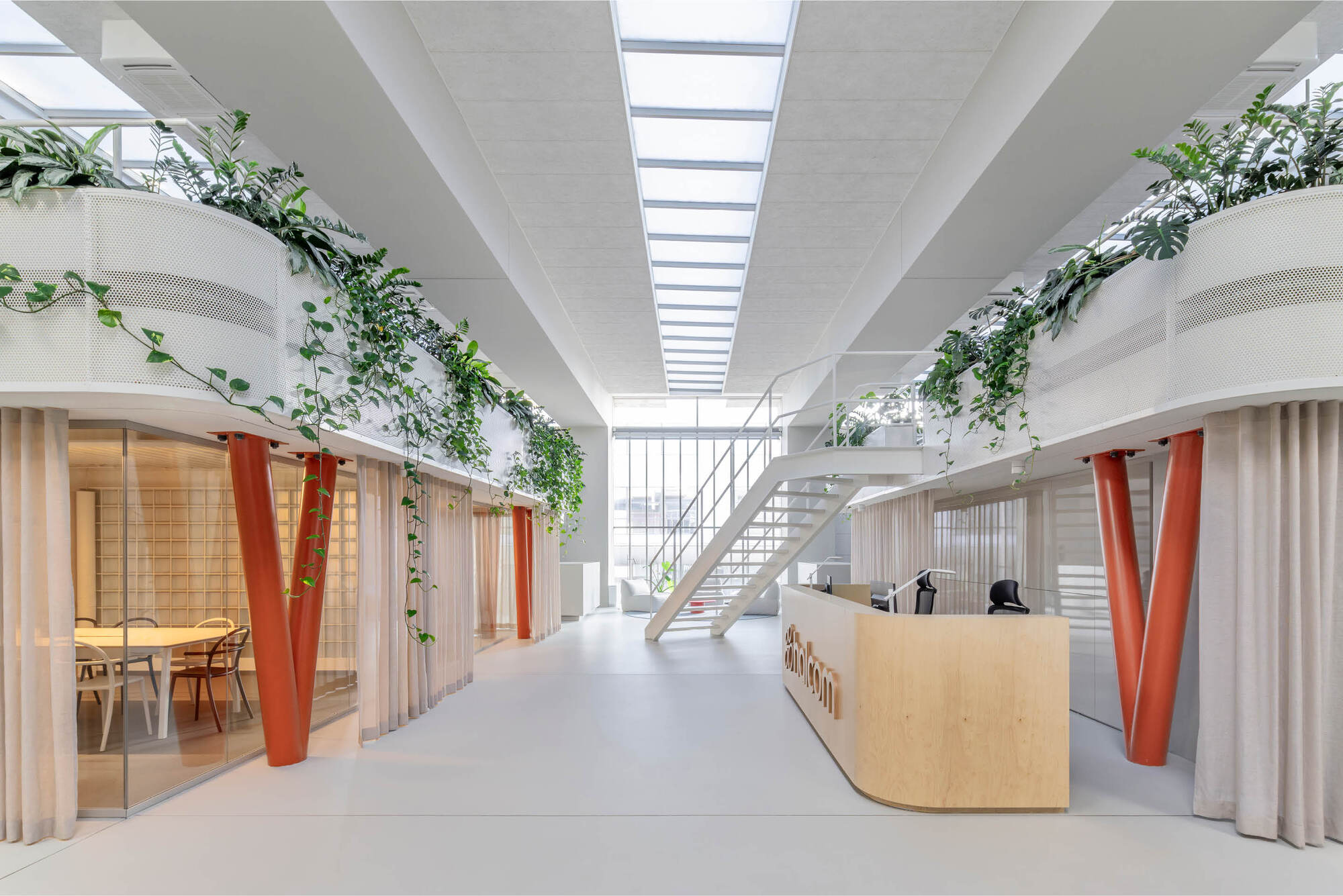 Central aisle between two planter-topped volumes, with orange V-columns, a white stair and a reception desk under a long skylight