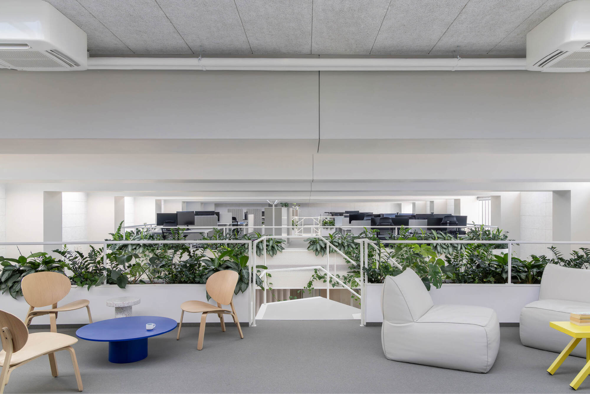 Upper-level lounge with wooden chairs, a blue round table and white pouf seats behind a planted balustrade overlooking the desks