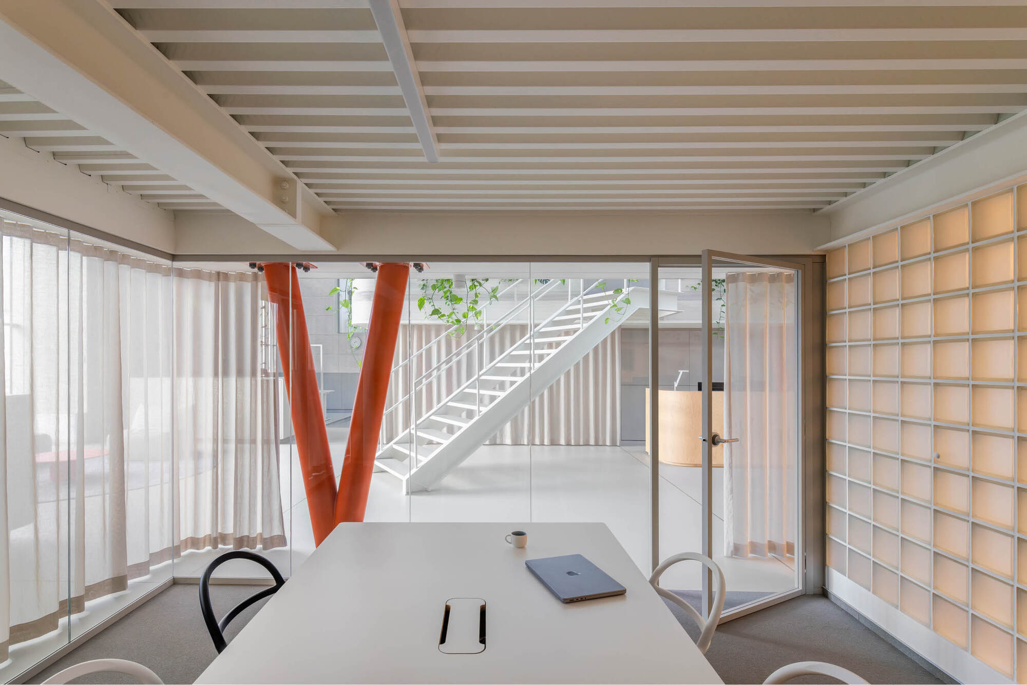 Meeting table seen through glass walls and sheer curtains, with an orange V-column, white stair and glass block wall glowing at the side