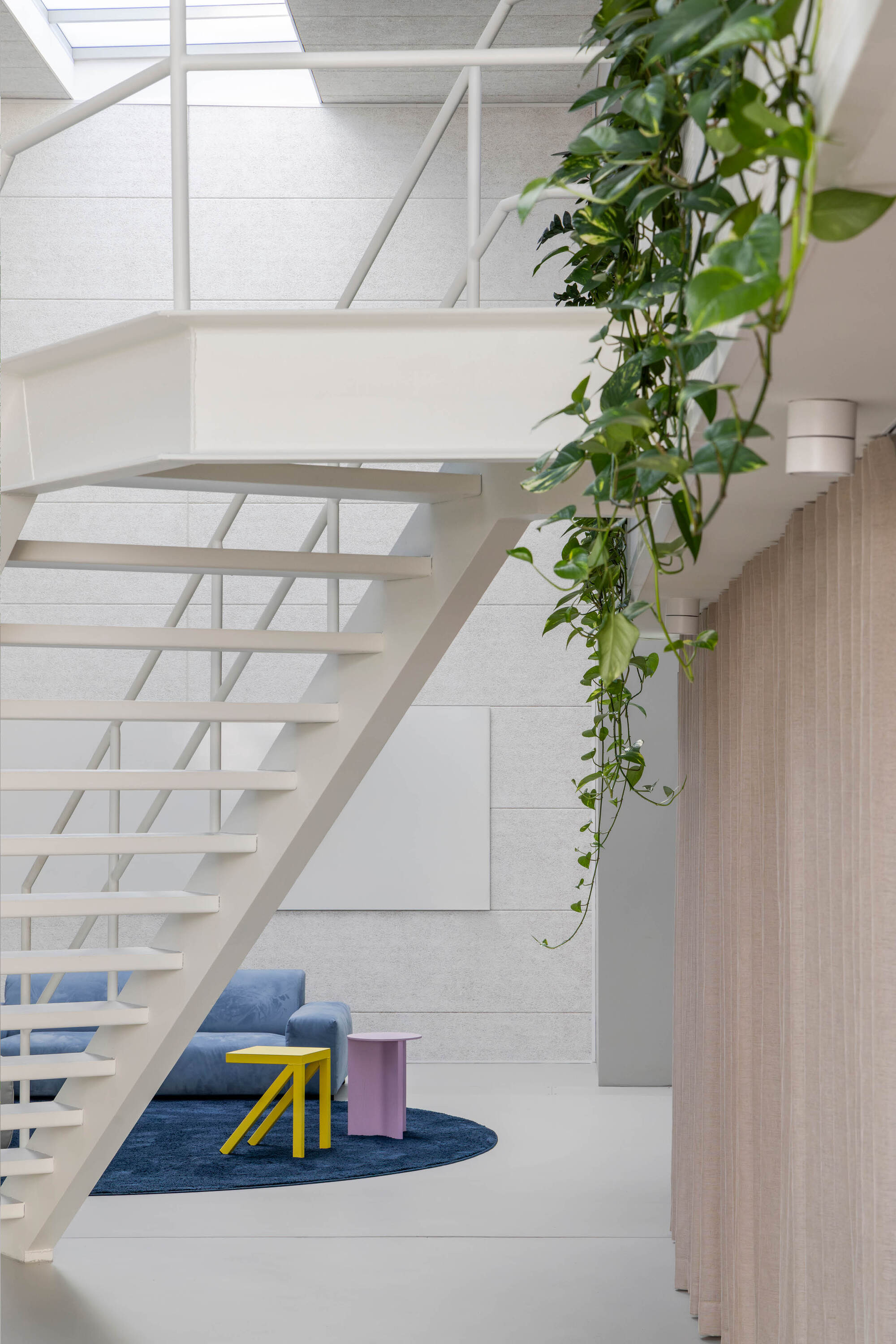 Underside of the white steel stair with a blue sofa, yellow stool and pink side table on a round rug