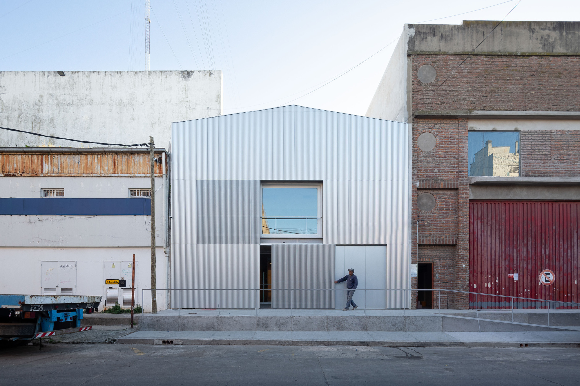 Gabled metal-clad facade with sliding corrugated panels between brick and plaster neighbors, a man walking past the open entrance in daylight