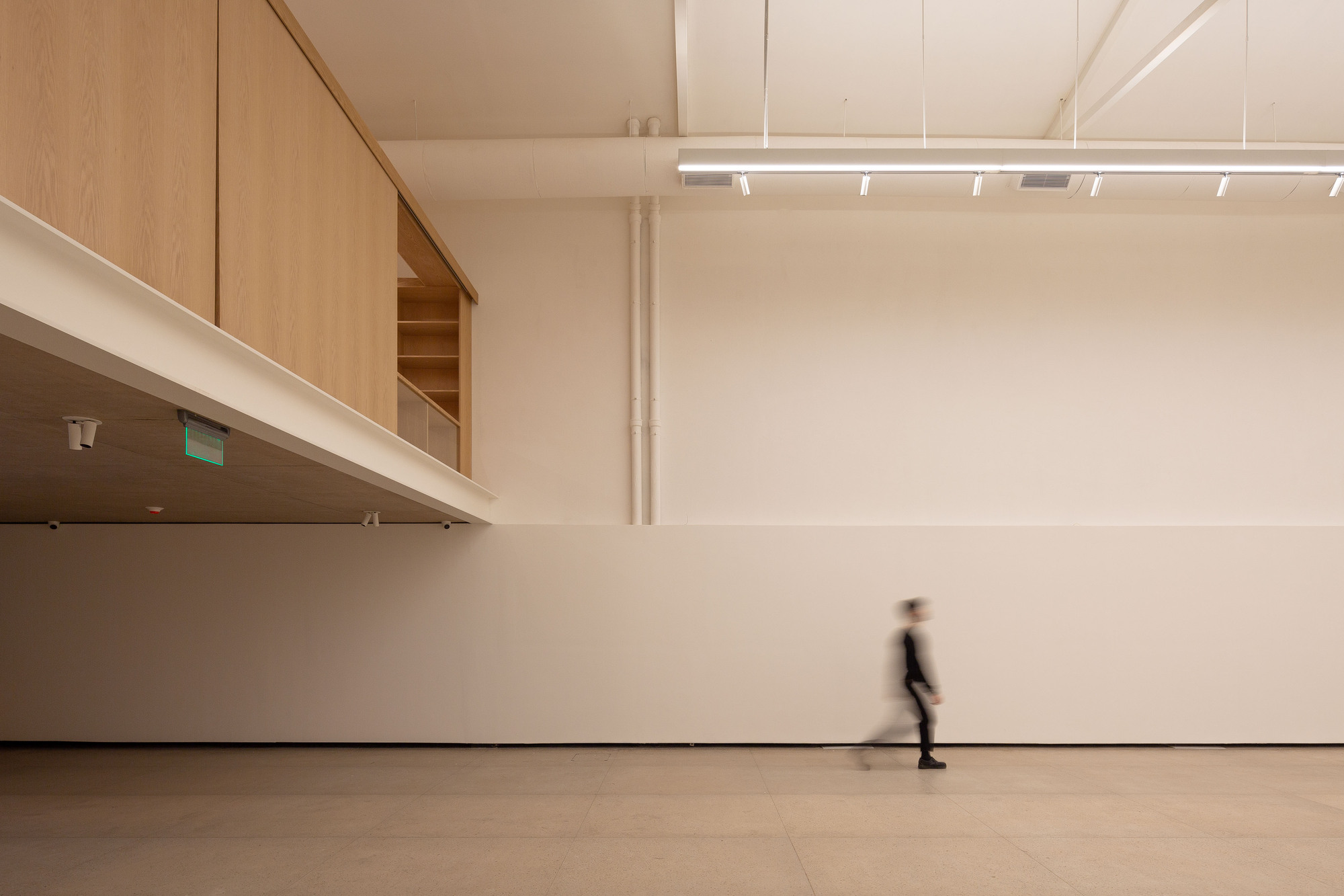 Gallery interior with a timber-clad mezzanine overhang and a blurred figure walking along a white wall under a linear light fixture