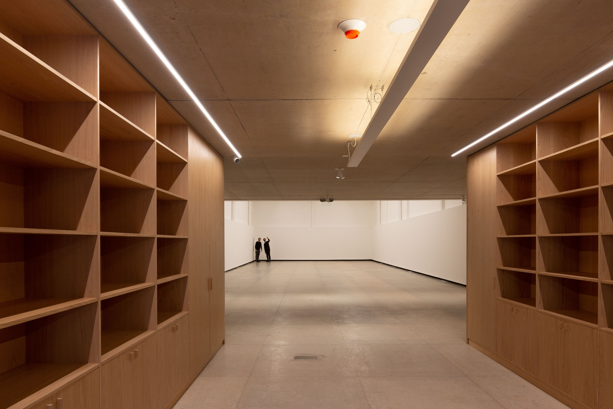 Corridor lined with oak shelving units on both sides, leading to a white exhibition room where two people stand under concrete ceilings