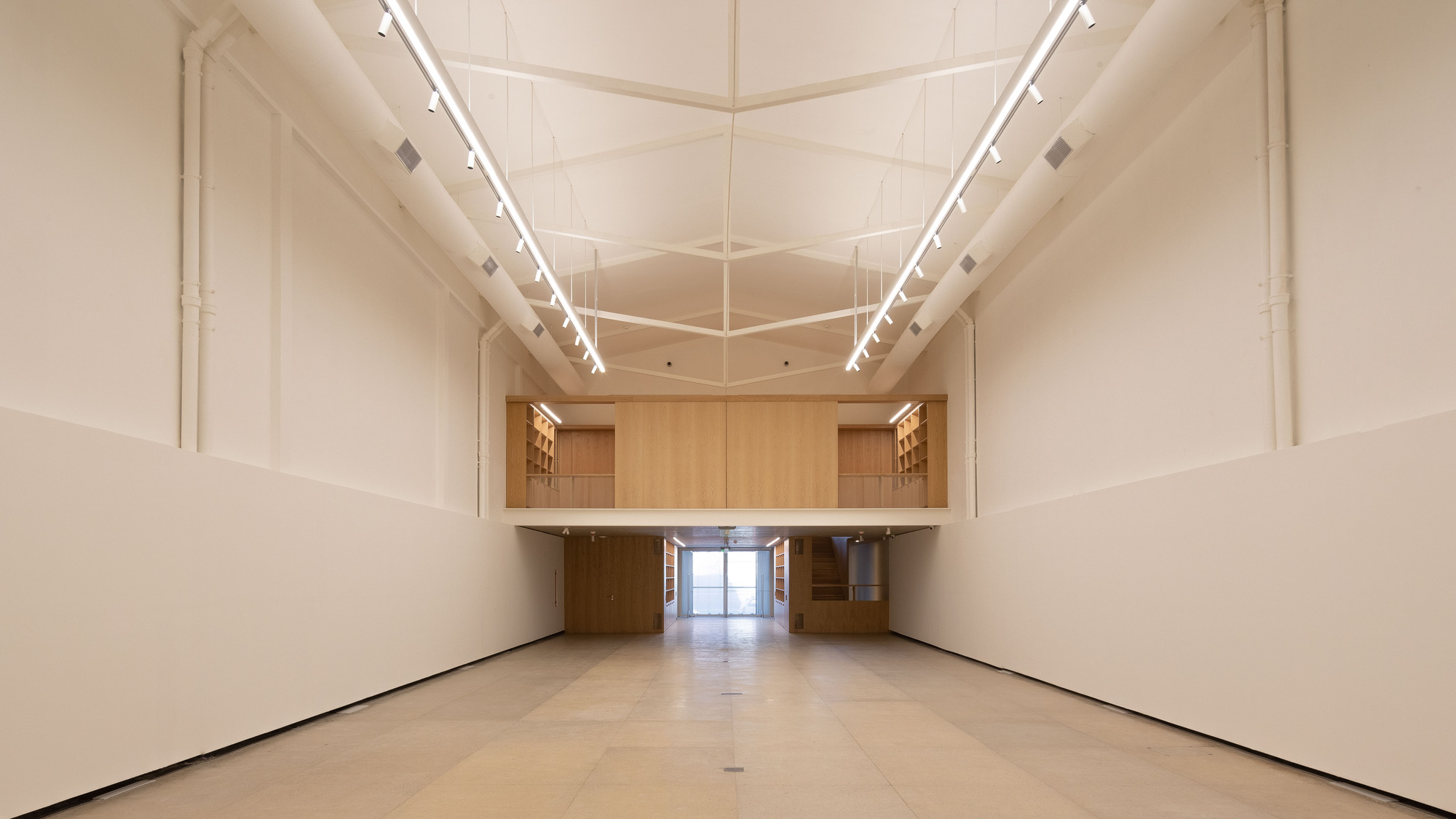 Long gallery hall with white walls, a timber mezzanine at the far end and linear lights hung below a ribbed ceiling