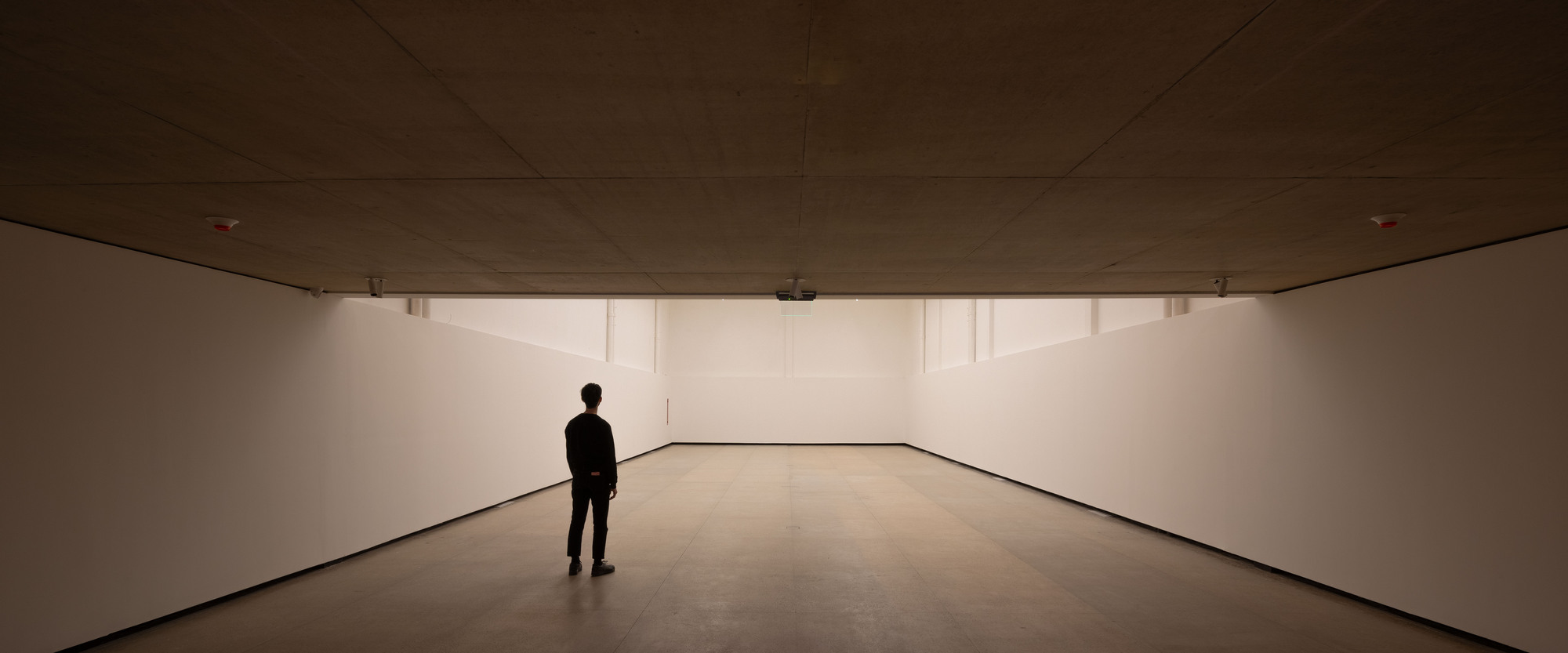Low-ceilinged white exhibition room with clerestory strips of daylight and a single figure standing on the pale stone floor
