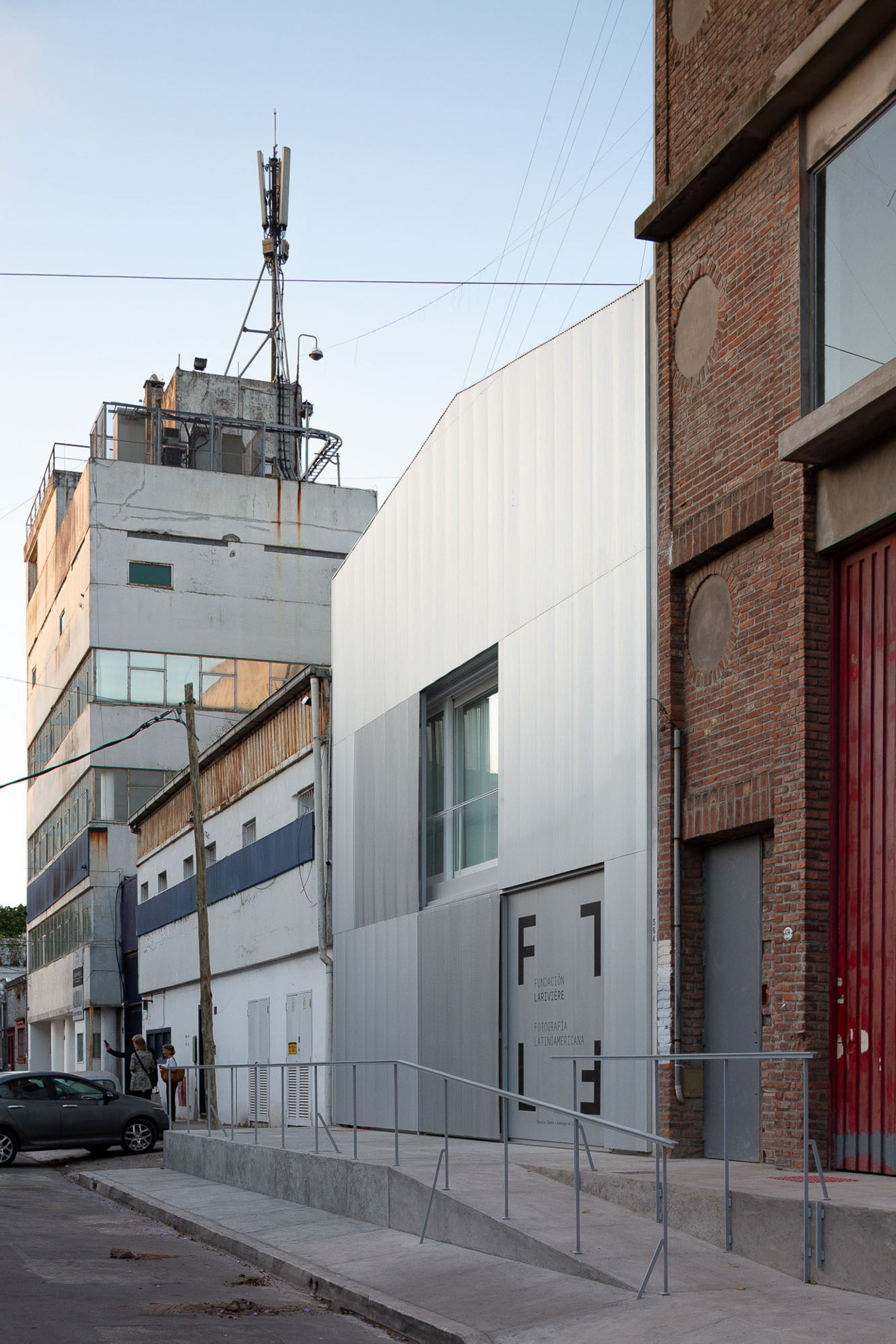 Corrugated metal clad gabled facade with a ramp and railings, squeezed between a red brick wall and older buildings at dusk