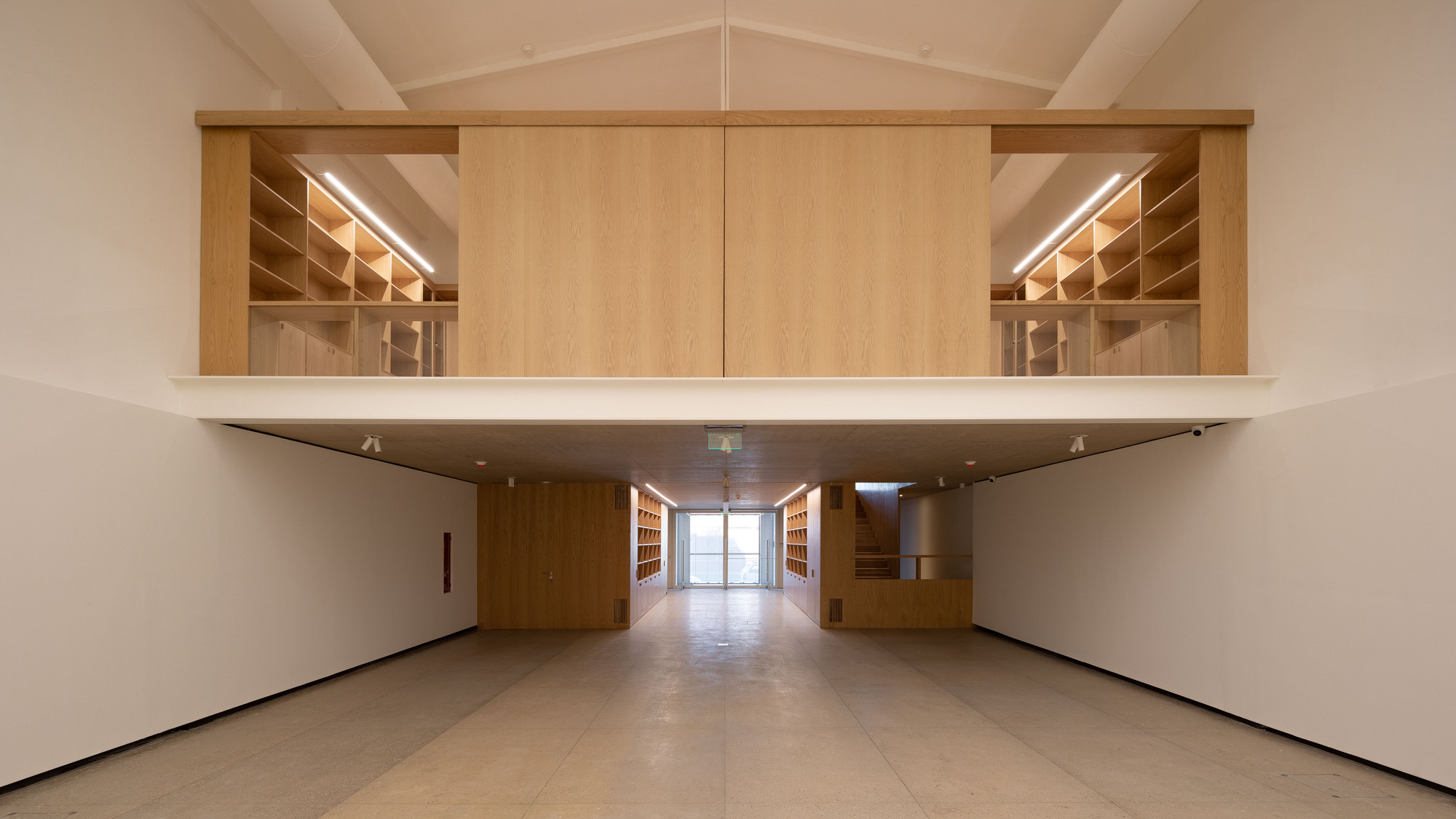 Gallery hall with an oak-clad mezzanine of open bookshelves floating above a concrete floor, lit by linear ceiling lights