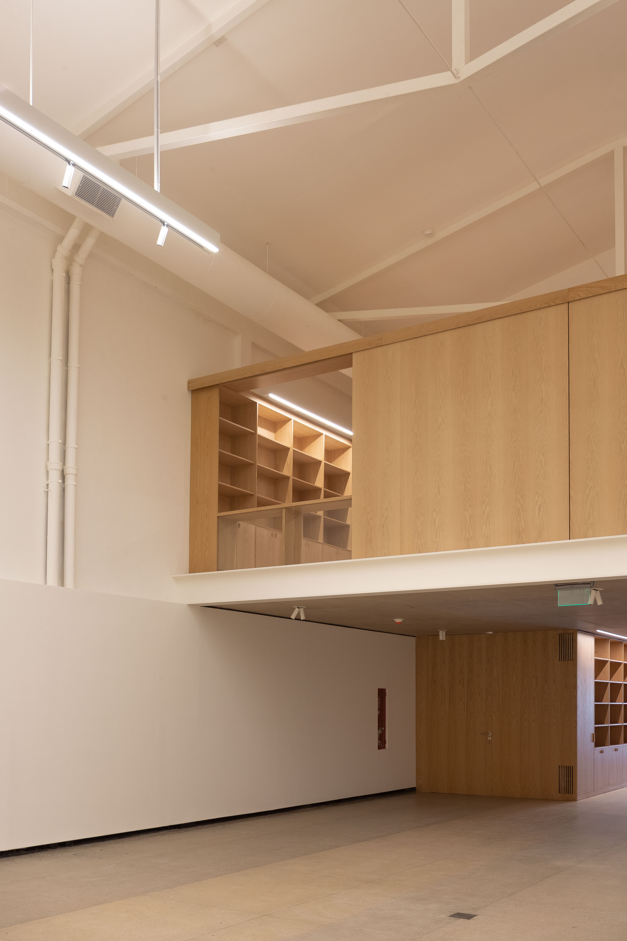 Corner view of the oak mezzanine box with shelving, white trusses and a suspended light fixture above the gallery