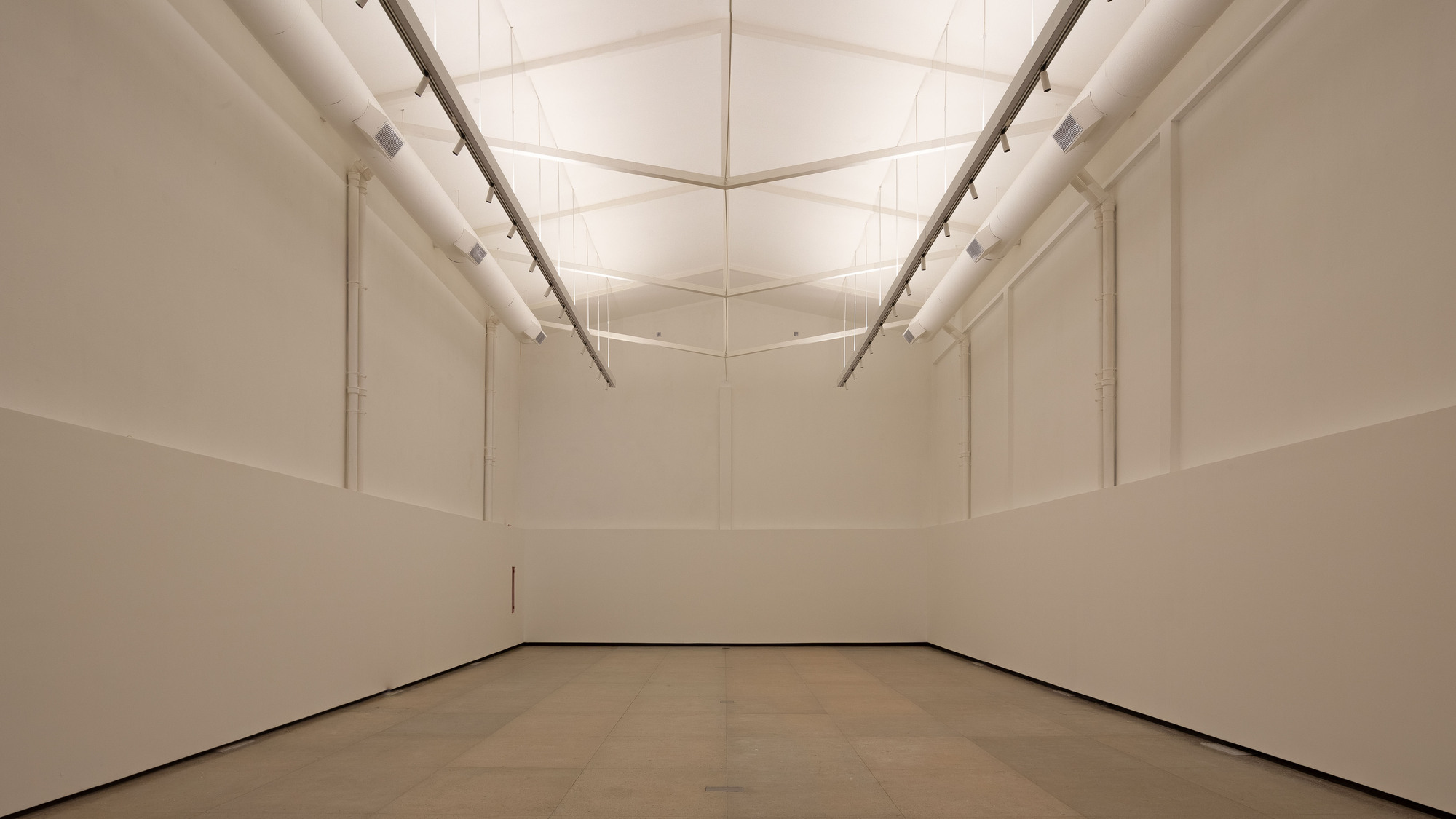 Empty white exhibition room with low partition walls, fabric ducts and a glowing translucent gabled ceiling
