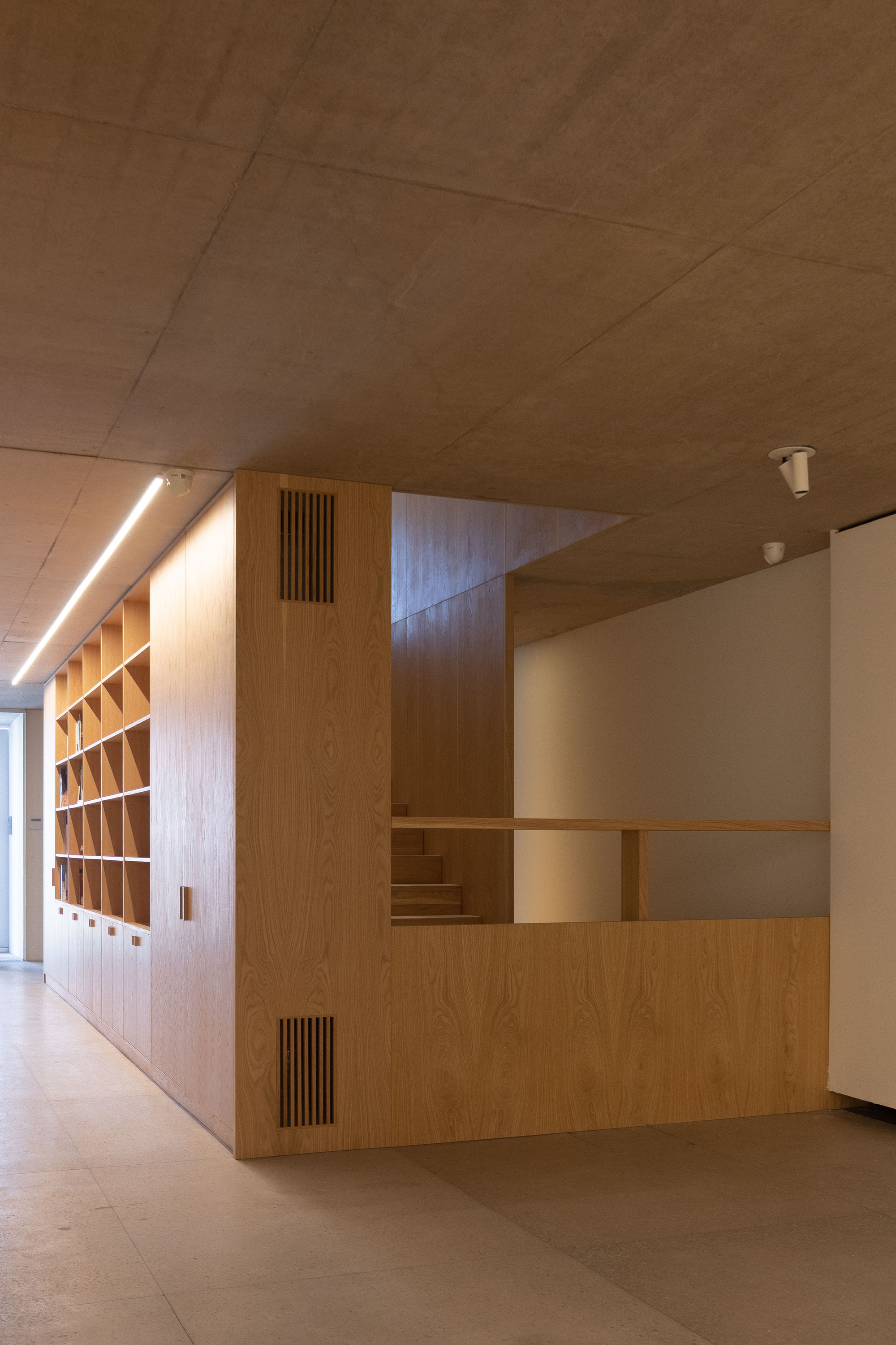 Oak joinery block with shelving, vent slots and a stair railing beneath a concrete panel ceiling and a track light