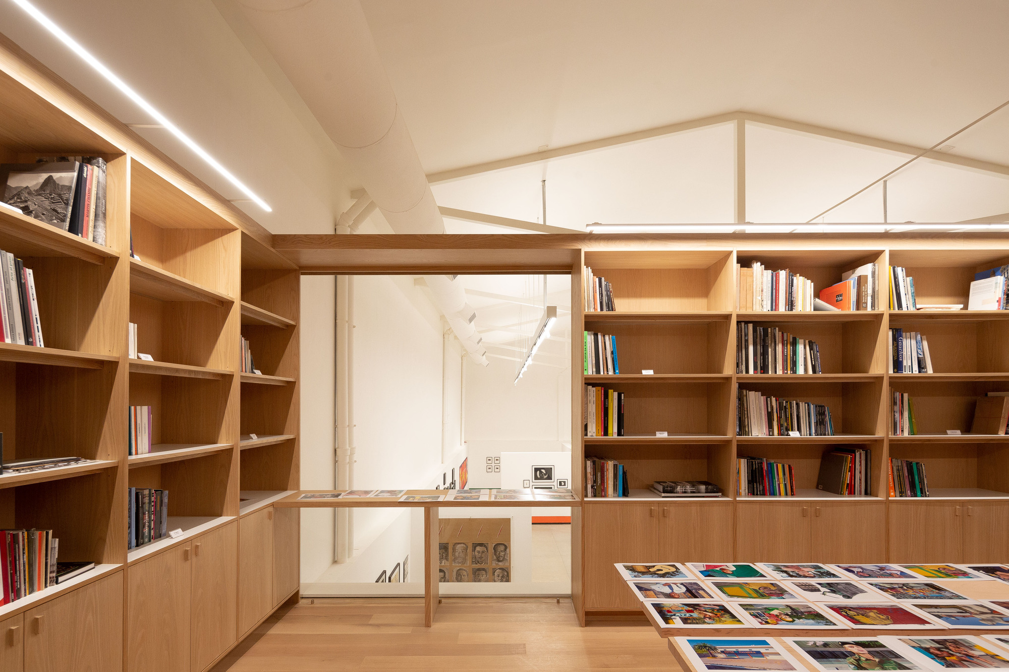 Upper-level library with oak shelves full of books, a table of printed photographs and a white truss roof beyond