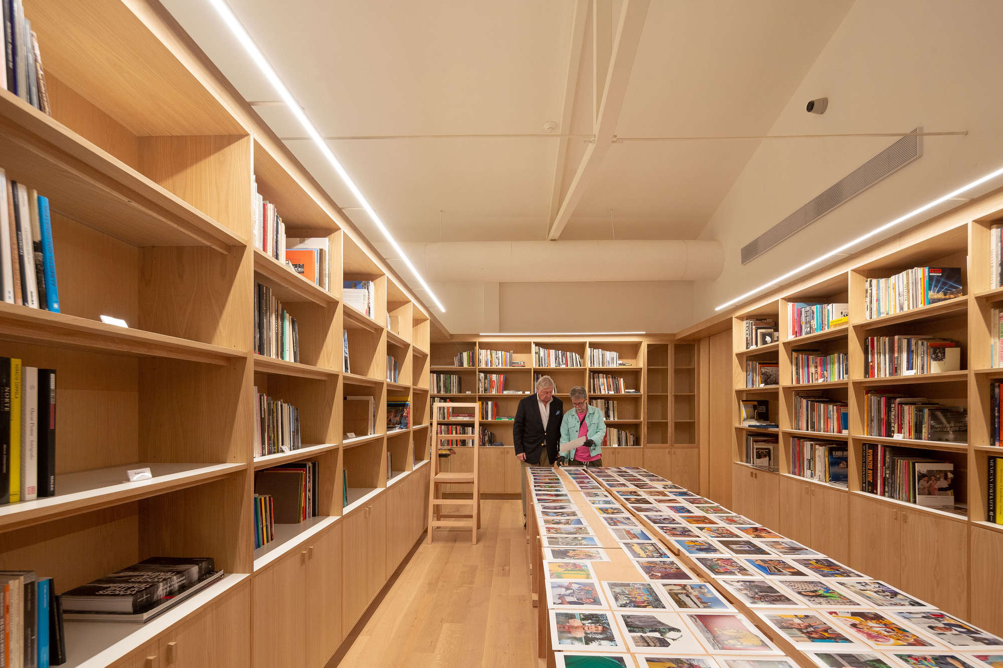 Oak bookshelves lining a long reading room where two visitors stand beside a table of laid-out prints under linear ceiling lights
