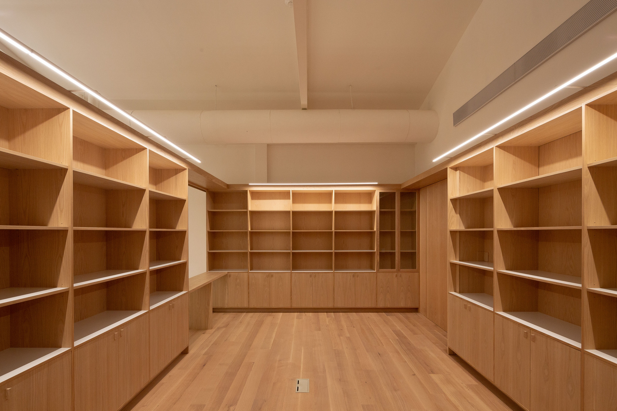 Empty oak shelving units and low cabinets along a room with a strip light and exposed round duct overhead