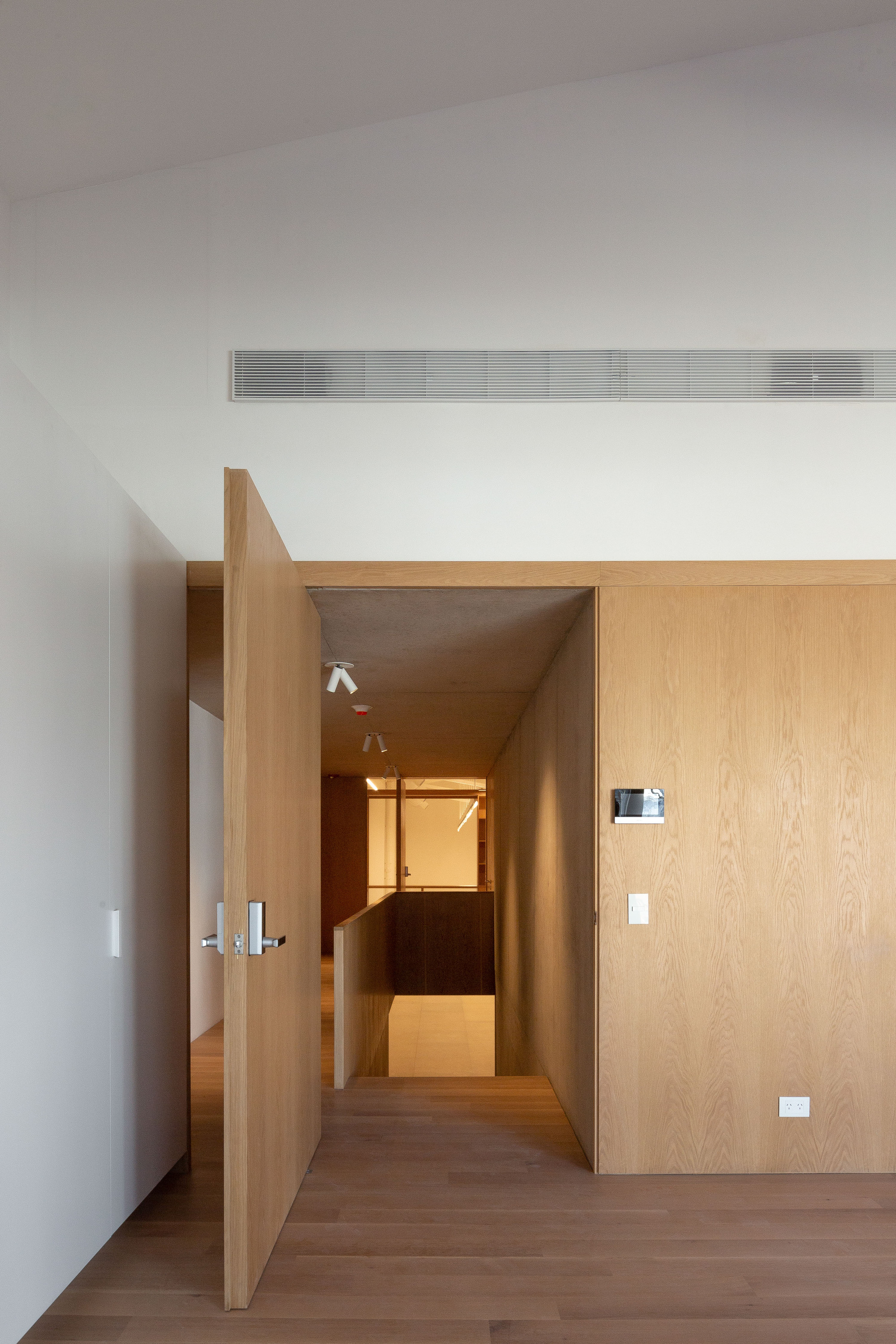 Open oak pivot door beside a wood-paneled wall, revealing a short passage toward a stair opening in soft daylight