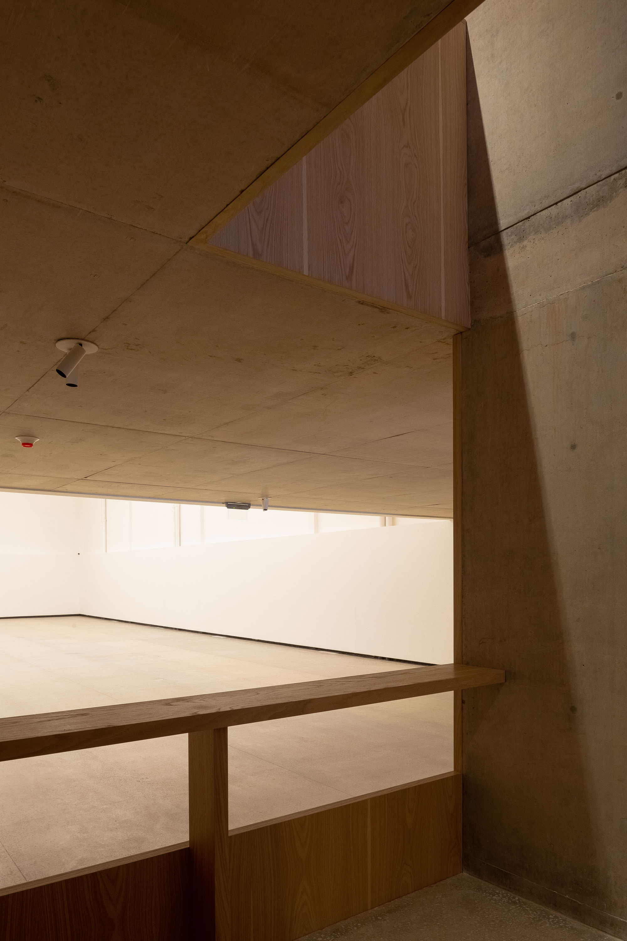 Oak railing and angled concrete wall framing a view into an empty white gallery lit by a clerestory band