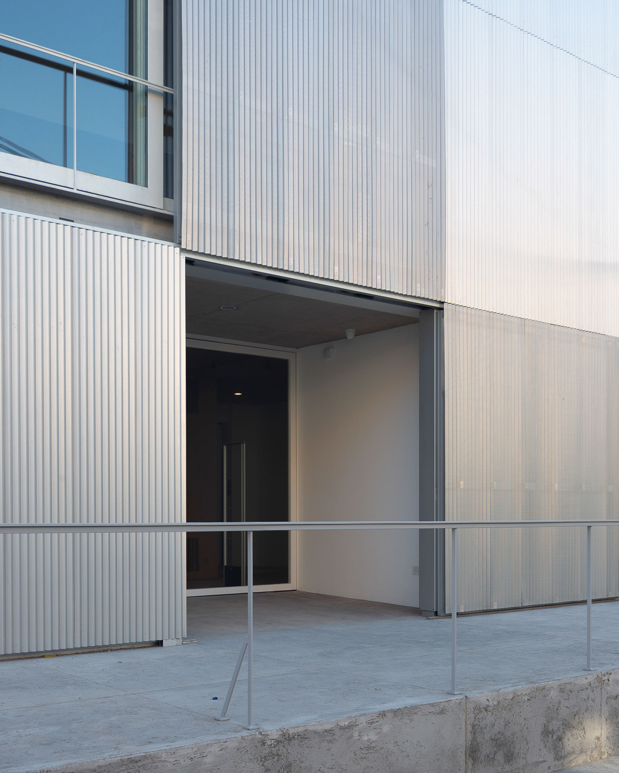 Recessed entrance in a corrugated aluminum facade, with a steel handrail along a concrete ramp at dusk