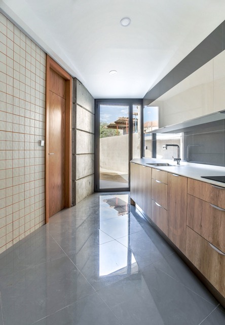 Narrow kitchen with oak cabinets, grid-tiled wall and glossy grey floor leading to a glazed door and daylight
