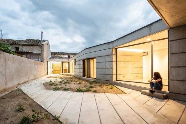 Courtyard at dusk with a concrete paved path, a seated woman and a glazed living room under cloudy sky