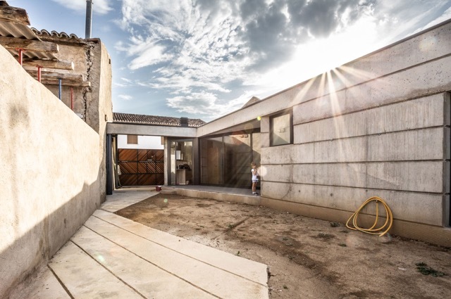 Sunburst over the courtyard where a concrete volume, hose coil and dirt ground meet a timber gate and a child