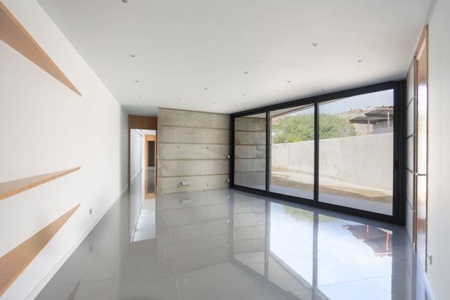 Living room with floor-to-ceiling black-framed sliding glazing, board-marked concrete wall and glossy grey tile reflecting daylight from a walled garden