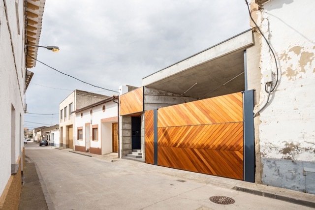 Street facade with a diagonal timber sliding gate beneath a concrete canopy, set between older whitewashed houses under an overcast sky