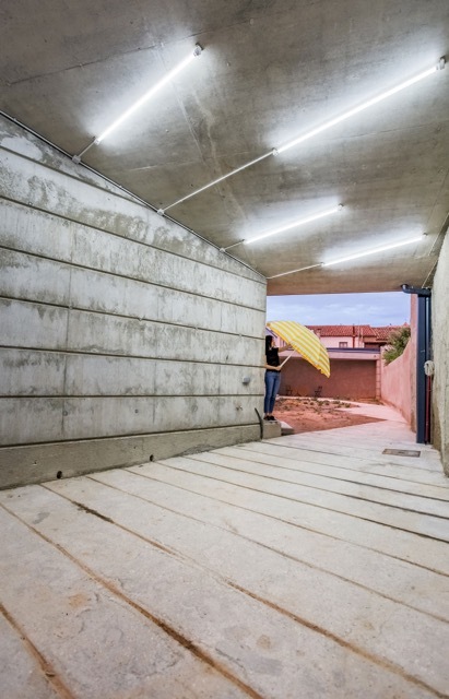 Covered concrete passage with board-marked walls and strip lights overhead, a person holding a yellow umbrella at the far opening at dusk