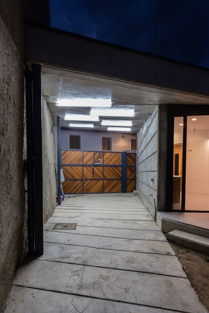 Night view along the concrete-paved passage toward the timber gate, with ceiling lights glowing and a glazed door at right