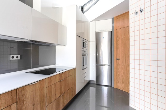 Galley kitchen with oak-fronted base cabinets, white countertop, steel fridge and a white tile wall with orange grout under a skylight