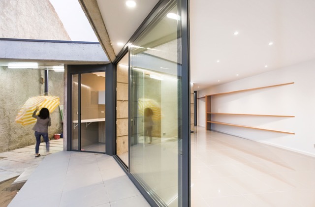 Corner of a glazed living space opening to a concrete patio where a woman walks with a yellow umbrella