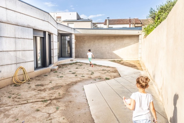 Two children run across a dirt courtyard and concrete paving beside a board-marked concrete wall in bright afternoon sun