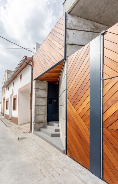 Street entrance with diagonal timber cladding, concrete block piers and grey steps leading to a dark door under cloudy skies