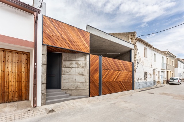 Street facade of diagonal timber panels and board-marked concrete, with a dark front door and a car parked down the lane