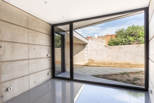 Interior corner with a board-marked concrete wall and a floor-to-ceiling glass wall facing a paved courtyard and leafy trees