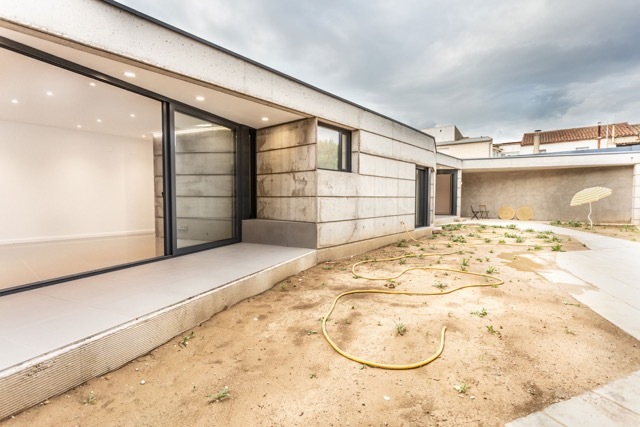 Board-formed concrete wall and sliding glass doors opening onto a sandy garden strewn with a yellow hose under an overcast sky