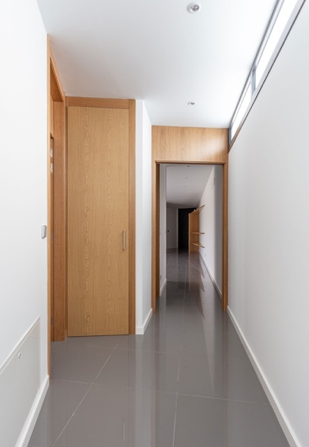 Narrow hallway with oak doors, white walls and glossy grey floor tiles, lit by a clerestory window along the right wall