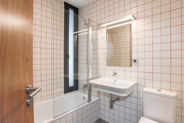 Bathroom with white tiles and rust-colored grout, a glass bath screen, wall-hung basin and slim vertical window beside an oak door