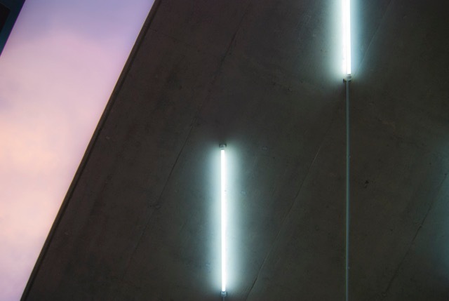 Two vertical fluorescent tubes mounted on a raw concrete wall that slants against a pink and violet dusk sky