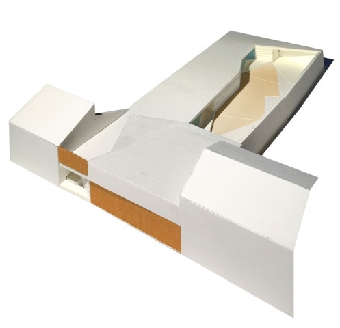White physical massing model with angled roof planes, orange wood-toned panels and a long open channel holding a tan sloped ramp, lit by soft daylight