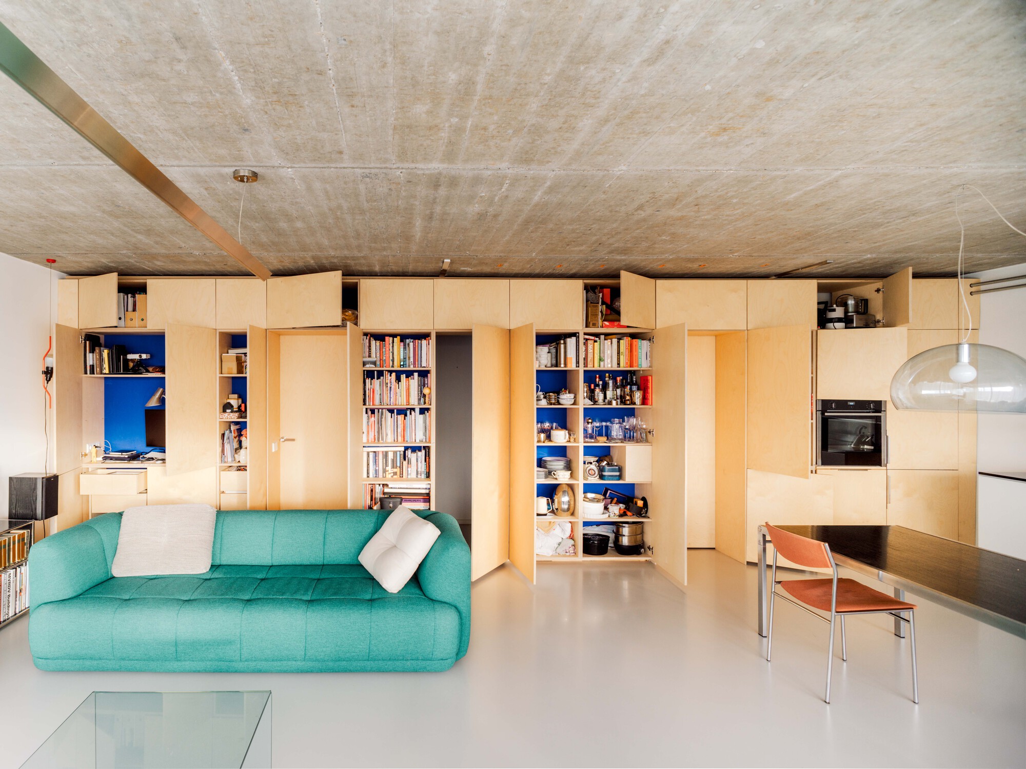 Birch plywood storage wall with open doors revealing bookshelves and kitchenware behind a teal sofa, under a board-marked concrete ceiling