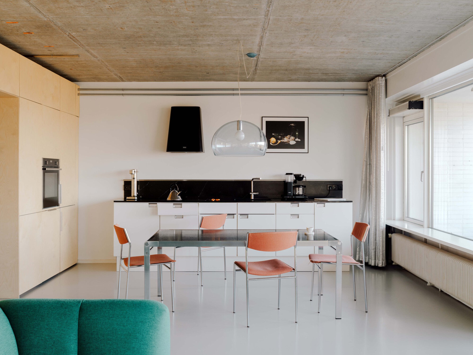 Open kitchen with white cabinets, black stone backsplash and a steel dining table with orange chairs beside a bright window
