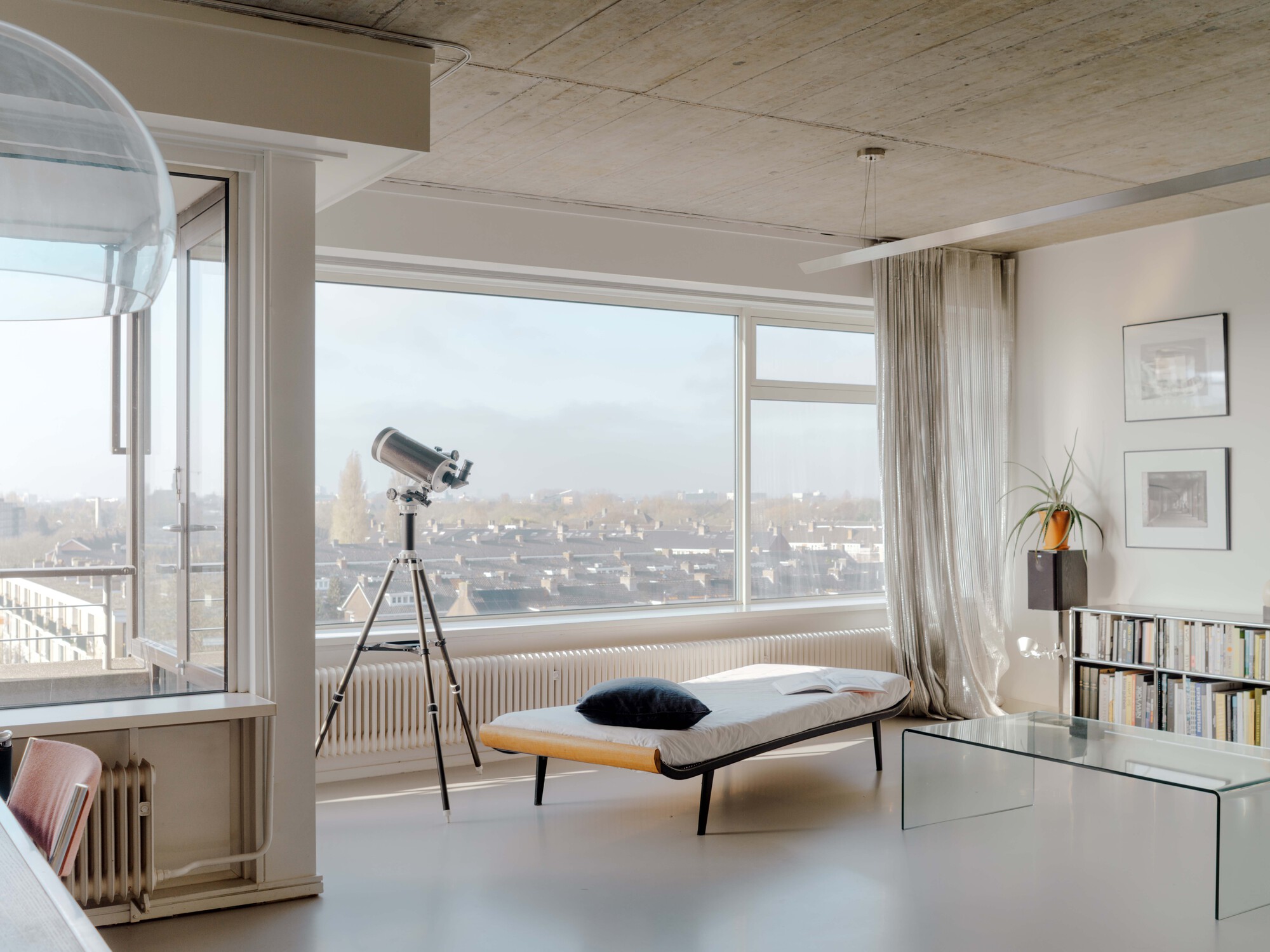 Daybed and telescope by a corner window overlooking rooftops, with sheer curtains and a glass coffee table in soft daylight