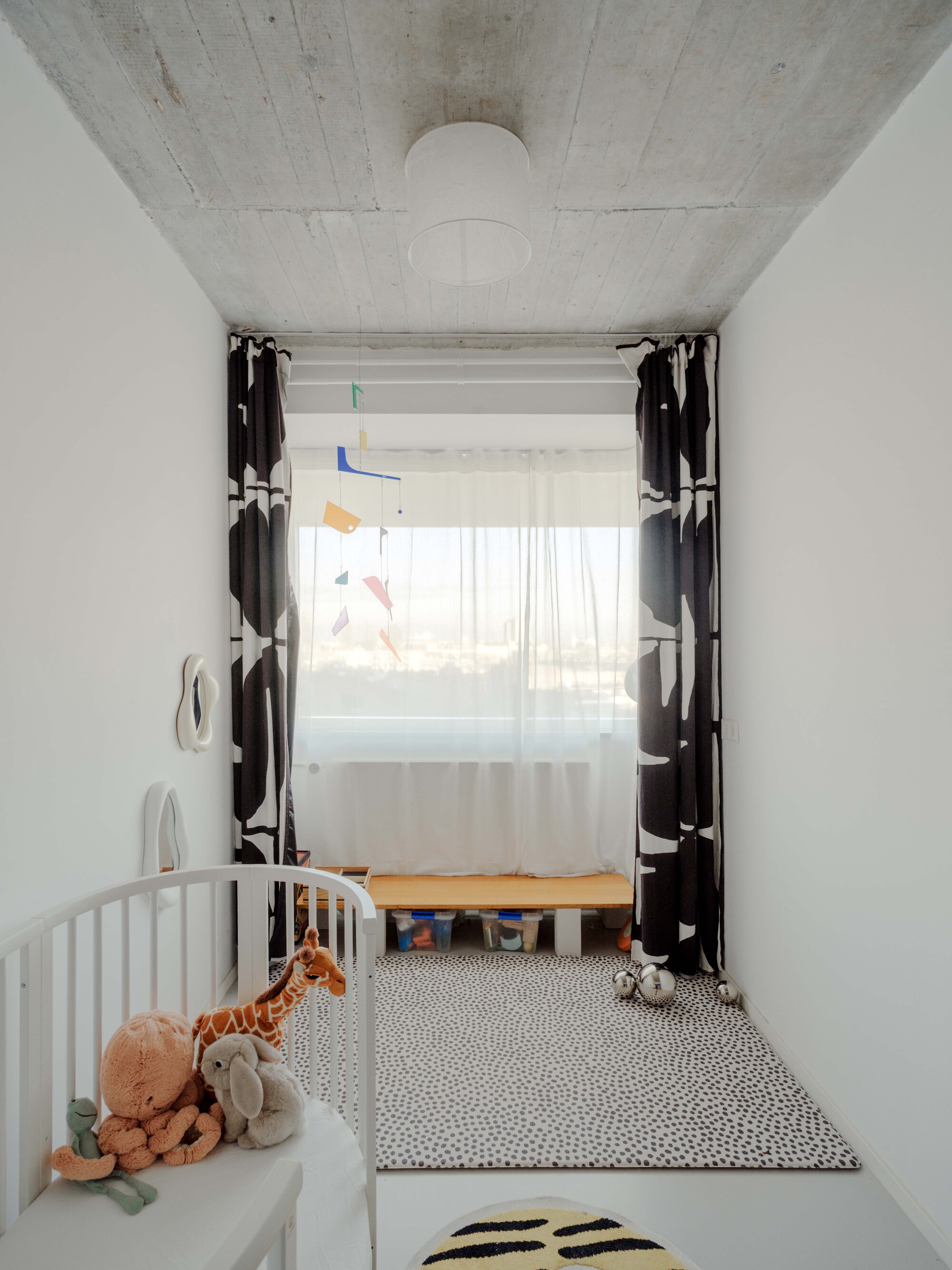 Narrow children's room with a white crib, spotted rug and black and white patterned curtains framing a hazy window