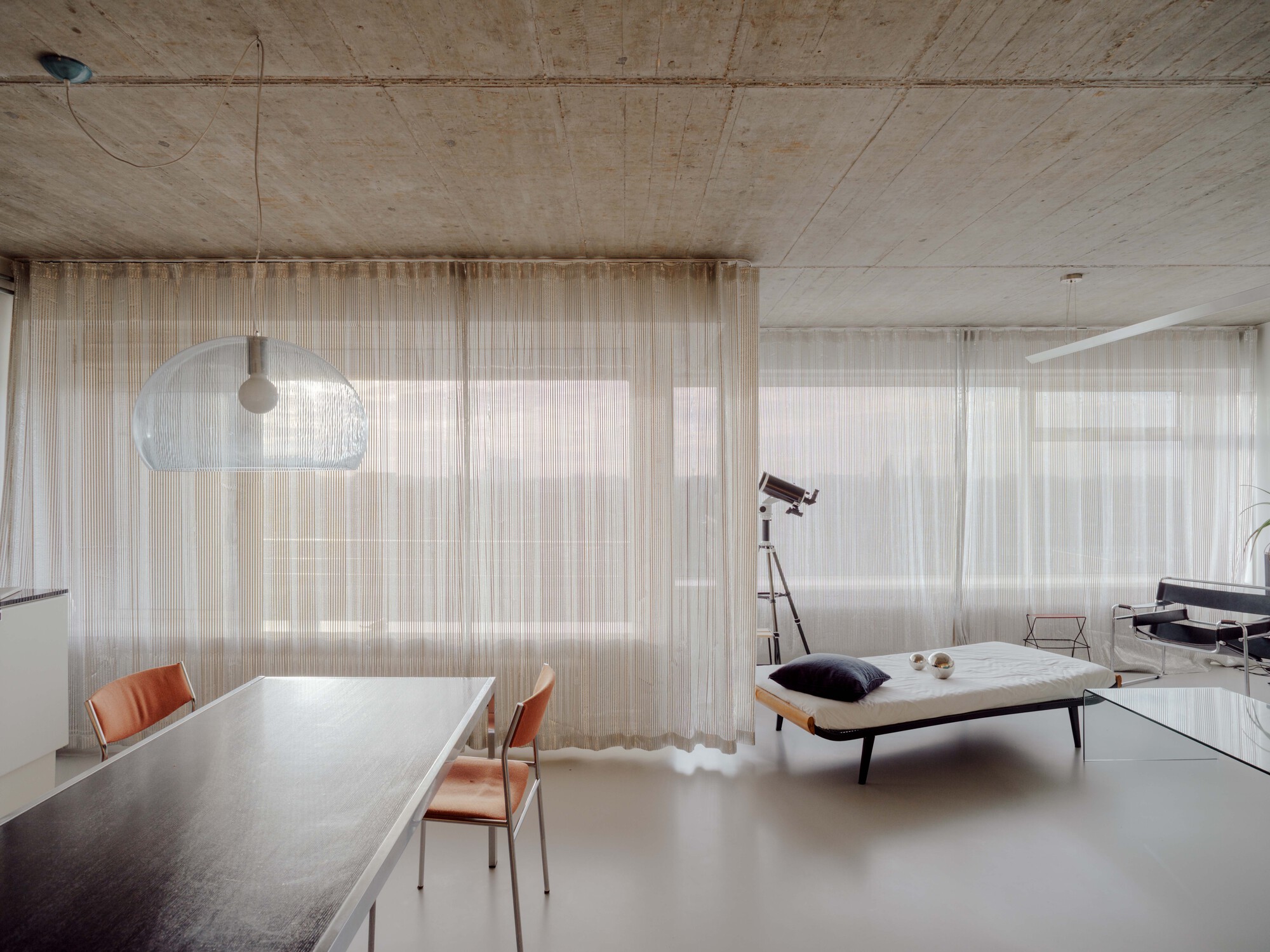 Long dark dining table and orange chairs facing floor-to-ceiling sheer curtains, with a daybed and telescope in diffuse light