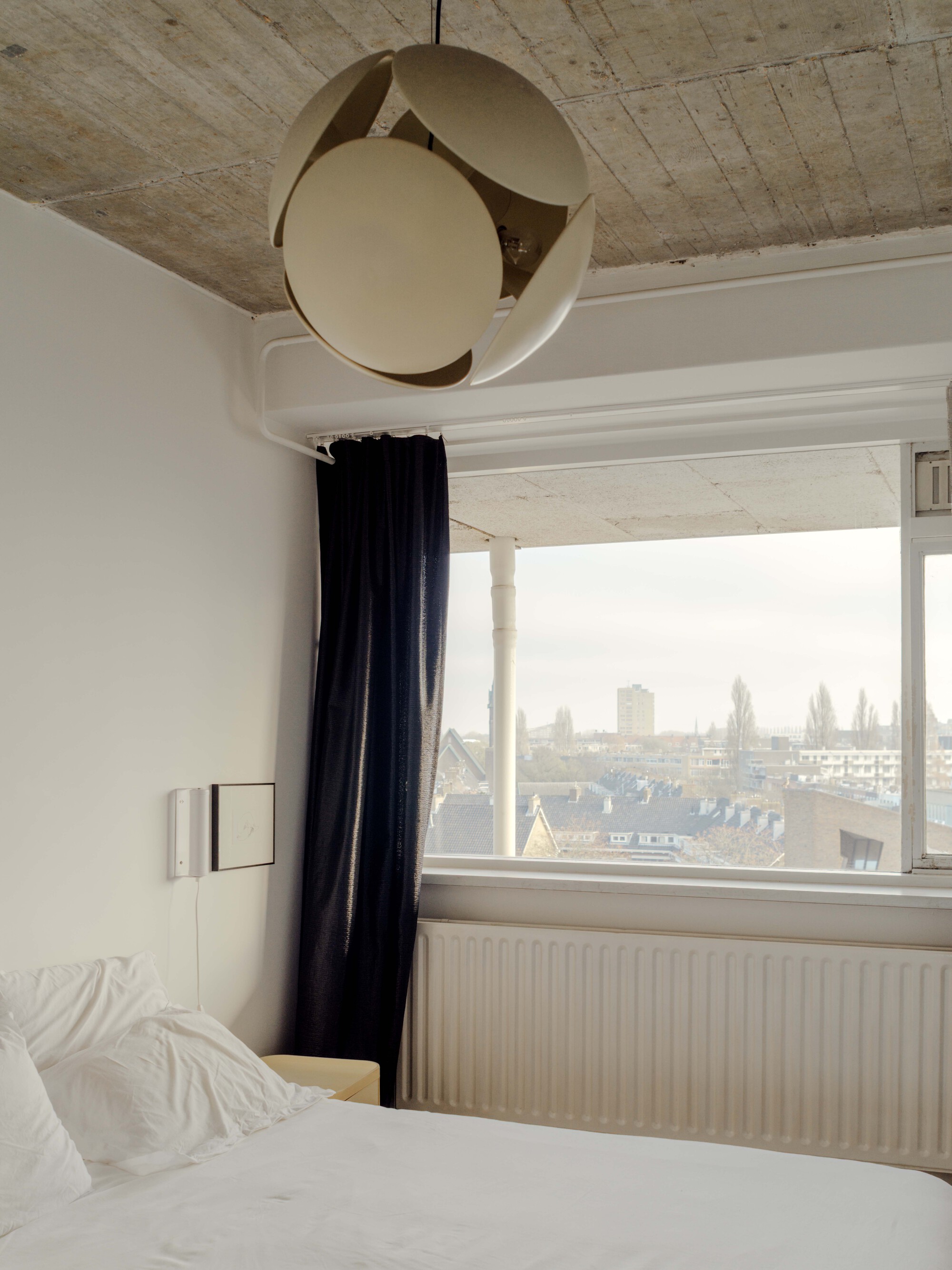 Bedroom corner with a round petal pendant lamp, navy curtain and radiator below a window onto rooftops on an overcast day