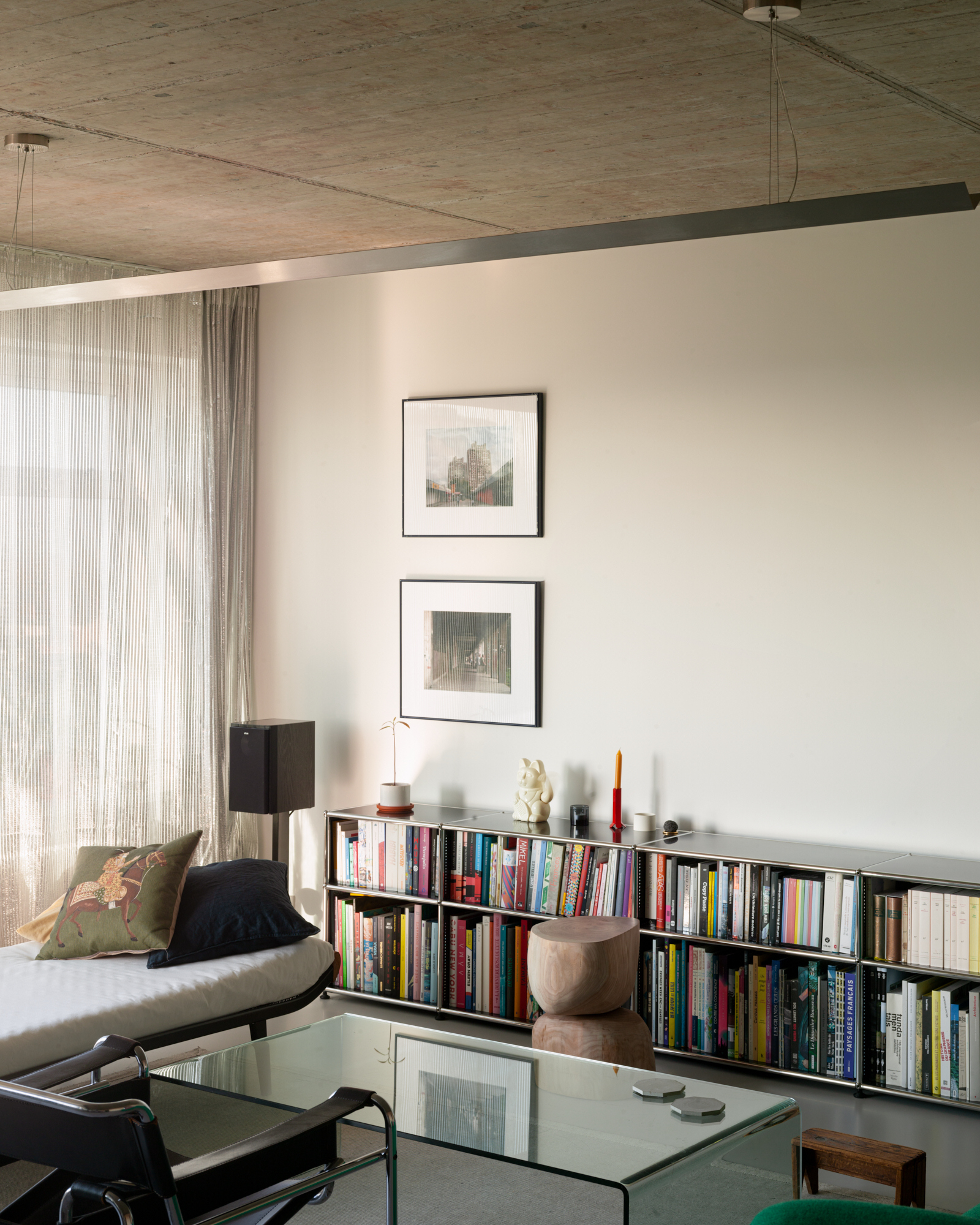 Low steel shelving of colorful books beneath two framed prints, with sheer curtains and a daybed in soft window light