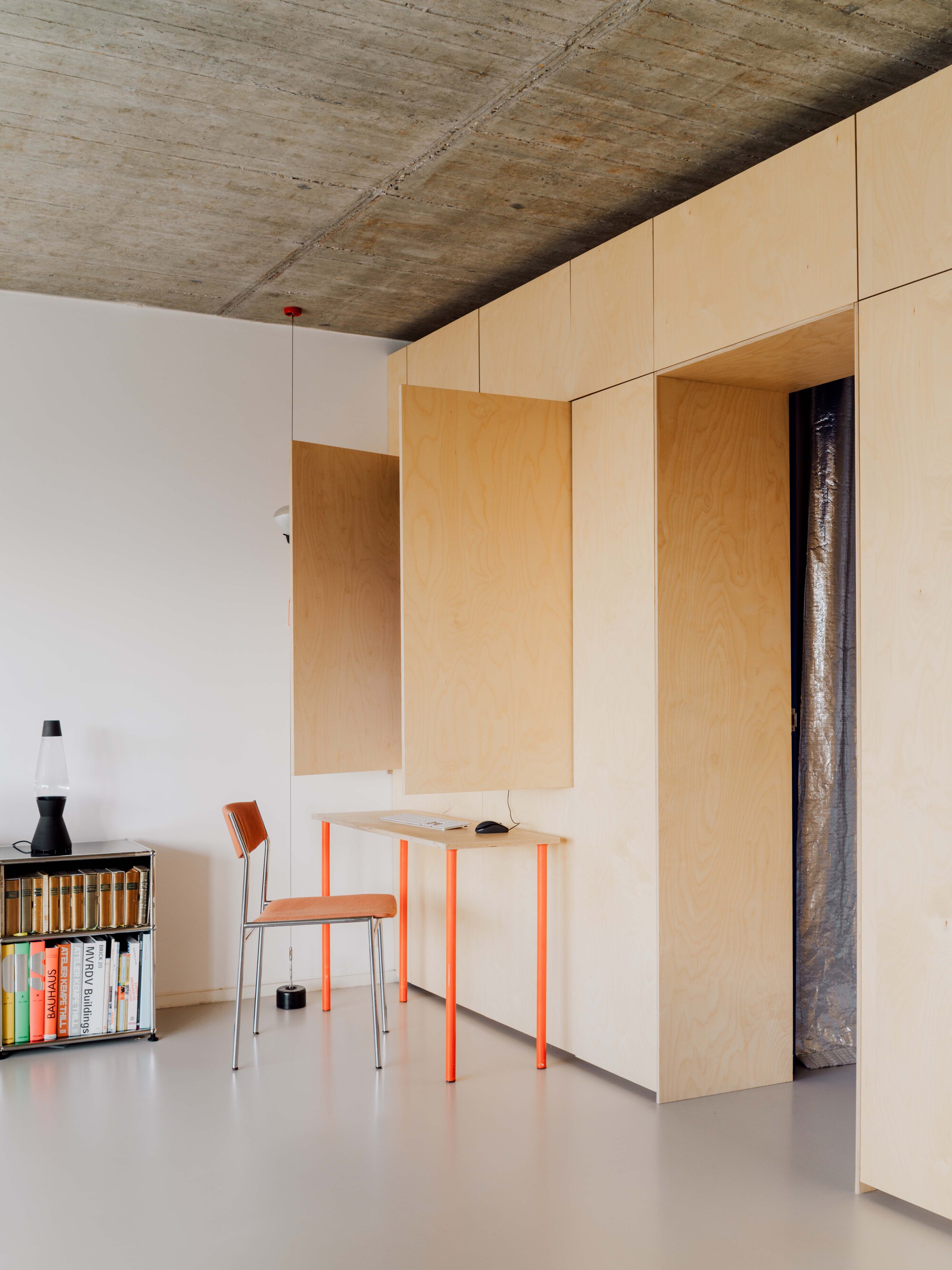 Birch plywood cabinet wall with a fold-down panel exposing a small desk, orange legs and a chair under a concrete ceiling