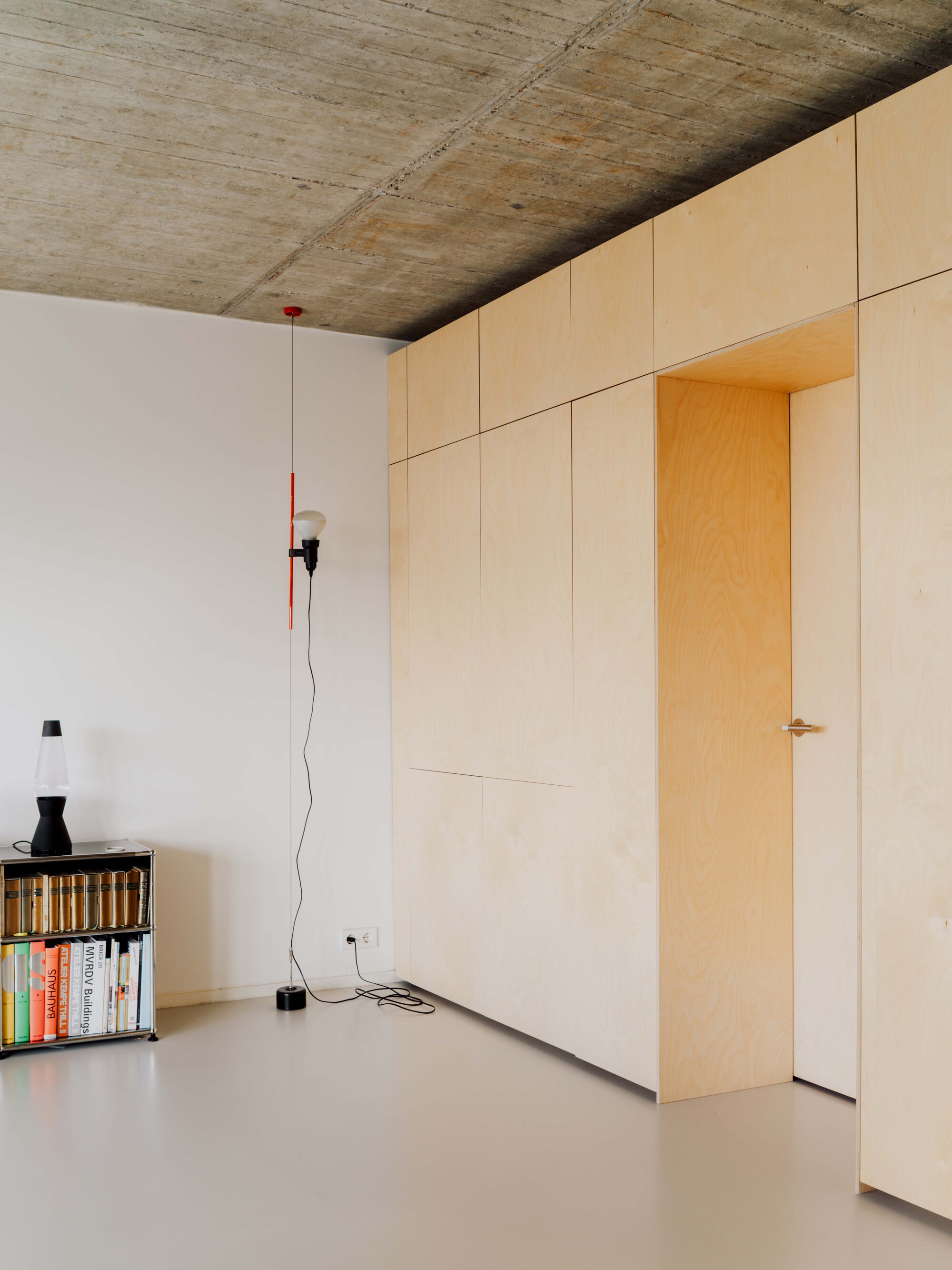 Closed plywood cabinet wall with a recessed doorway and a red-stemmed floor lamp beside a small bookshelf
